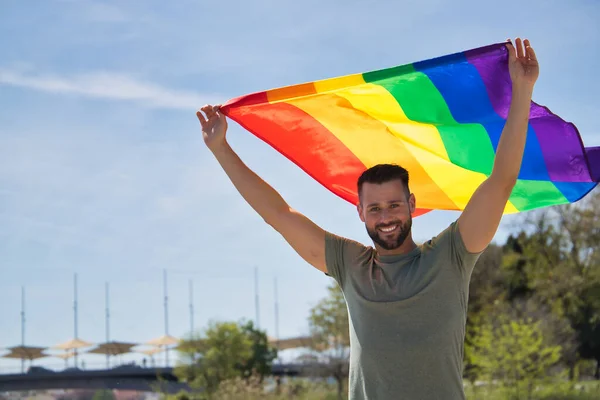 stock image Young and handsome gay man, with a beard and green shirt, with blue eyes, perfect smile and waving a gay pride flag, smiling. Concept of gay pride, homosexual, lgtbi, pride day.