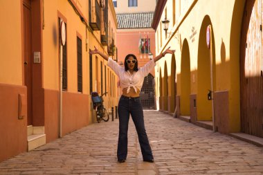 Young and beautiful South American woman in white knotted shirt, jeans and sunglasses with open arms in the middle of a cobblestone street. Concept beauty, fashion, vacation, travel, urban, city.