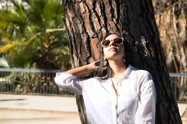 若く美しい女性 南アメリカ人は サングラスと白いシャツを着て 彼女の髪に触れながら太陽の光線を取る木の幹にもたれています コンセプト旅行 美しさ ファッション — ストック写真