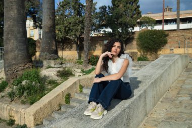 Genç ve güzel bir kadın, esmer, kıvırcık saçlı, sevecen bir jest, beyaz gömlek ve kot pantolon giymiş, beton bir duvarda oturuyor. Güzelliği, modayı, modeli, sevgiyi, eğilimi kavra.