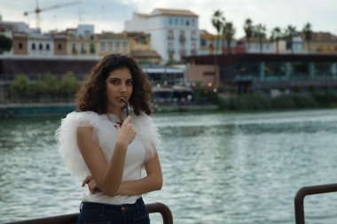 Genç ve güzel bir kadın, esmer, kıvırcık saçlı, güzel bir gülümsemesi olan, dişlerinin arasına beyaz bir gömlek, kot pantolon ve güneş gözlüğü takan baştan çıkarıcı bir tavırla. Güzellik, moda, baştan çıkarma kavramları.