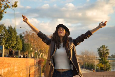 Genç ve güzel bir kadın, esmer, kıvırcık saçlı, deri ceketli, siyah bereli, kolları açık ve gözleri kapalı. Güzelliği, modayı, modeli, eğilimi, barışı, özgürlüğü kavra.