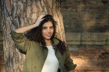 Genç ve güzel bir kadının portresi, esmer, kıvırcık saçlı, yeşil ceketli ve beyaz gömlekli, bir ağacın gövdesine yaslanmış ve baştan çıkarıcı bakışlı. Güzellik, moda, baştan çıkarma kavramları.