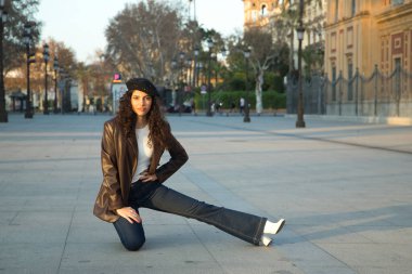 Genç ve güzel bir kadın, esmer, kıvırcık saçlı, deri ceketli ve siyah bereli, şehri ziyaret ederken yerde poz veriyor. Güzellik, moda, trend, seyahat, tatil kavramları.