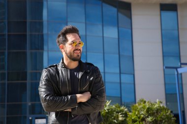 Sakallı, güneş gözlüklü, deri ceketli, kot pantolonlu, kollarını kavuşturmuş, mutlu bir şekilde gülümseyen yakışıklı bir genç. Güzellik, moda, trend, modern, başarılı, mutluluk kavramı.