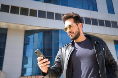Sakallı, güneş gözlüklü, deri ceketli, cep telefonundan sosyal ağları kontrol eden yakışıklı bir genç. Güzelliği, modayı, eğilimi, uygulamayı, akıllı telefonu, sosyal ağları kavra.