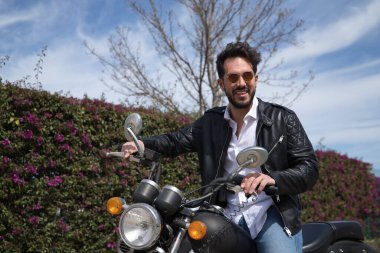Sakallı, güneş gözlüklü, deri ceketli, beyaz gömlekli ve kot pantolonlu, büyük yer değiştiren motosikletli yakışıklı bir genç. Güzellik, moda, trend, motor, motosiklet, çıplak kavram.