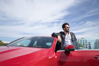 Sakallı, güneş gözlüklü, deri ceketli, beyaz gömlekli, kırmızı spor arabasının çatısına yaslanmış yakışıklı bir genç. Güzellik, moda, trend, lüks, motor, spor kavramı.