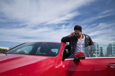 Sakallı, güneş gözlüklü, deri ceketli, beyaz gömlekli, kırmızı spor arabasının çatısına dayanan, gözlüğünü takan yakışıklı bir genç. Güzellik, moda, trend, lüks, motor, spor kavramı.