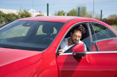 Kırmızı spor arabasının içinde sakallı, güneş gözlüklü ve beyaz gömlekli yakışıklı bir genç. Güzellik, moda, trend, lüks, motor, spor, kazanan.