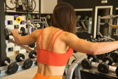 Genç ve güzel bir kadın spor salonundaki aynaya yansıyan dambıllarla omuz egzersizleri yapıyor. Fitness konsepti, ağırlık, vücut geliştirme, jimnastik, kaslar.