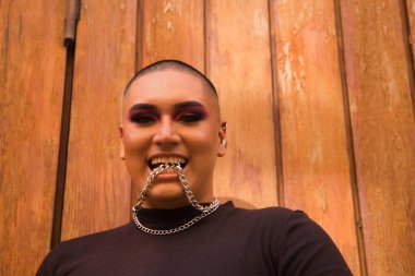 İkili olmayan bir kişinin portresi, genç ve Güney Amerikalı, arka planda tahta bir duvar bulunan dişli metal bir zinciri ısırıyor. Kavram kraliçesi, lgbtq +, gurur, nonoş.