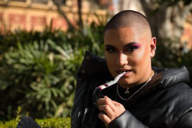 İkili olmayanların portresi, genç ve Güney Amerikalı, ağır makyajlı, dişleriyle ısıran güneş gözlüğü tapınağı ve bitki örtüsüyle çevrili. Kavram kraliçesi, lgbtq +, gurur, nonoş.