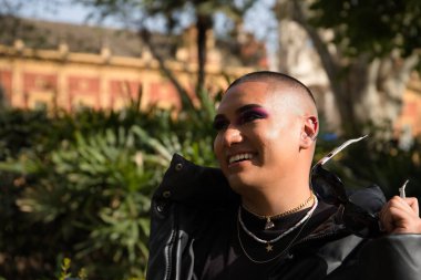 İkili olmayan bir kişinin portresi, genç ve Güney Amerikalı, ağır makyajlı, elinde güneş gözlükleriyle gülümseyen ve yeşilliklerle çevrili. Kavram kraliçesi, lgbtq +, gurur, nonoş.