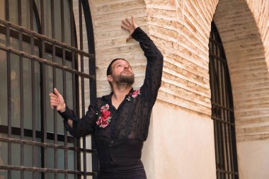 Sakallı, at kuyruklu, siyah şeffaf tişörtlü, siyah puantiyeli, kırmızı güllü, siyah pantolonlu ve ceketli, pencere ızgarasına yapışmış flamenko dansı yapan genç bir adam. Sanat kavramı, dans..