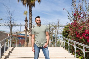 Sakallı, yeşil tişörtlü ve kot pantolonlu, parkın merdivenlerinde poz veren yakışıklı bir genç. Güzellik kavramı, moda, moda, model, modern.