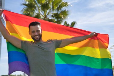 Genç ve yakışıklı eşcinsel, sakallı ve yeşil gömlekli, mavi gözlü, mükemmel gülüşlü ve elinde gay gurur bayrağı olan, gülümseyen. Gay gururu kavramı, homoseksüel, lgtbi, gurur günü.