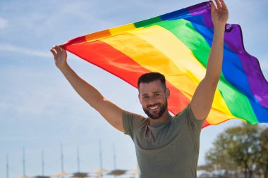 Genç ve yakışıklı eşcinsel, sakallı ve yeşil gömlekli, mavi gözlü, mükemmel gülüşlü ve gay gurur bayrağı sallayan, gülümseyen. Gay gururu kavramı, homoseksüel, lgtbi, gurur günü.