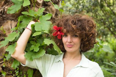 Portrait of mature and attractive woman with curly brown hair and a red flower in the ear, leaning on a palm tree with green leaves. Spring concept, flowers, beauty, hairstyle, curls.