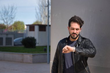 Sakallı, deri ceketli, lüks kol saatine bakan yakışıklı bir genç. Güzellik, moda, trend, modern, lüks, mücevher, zaman, saat kavramı.