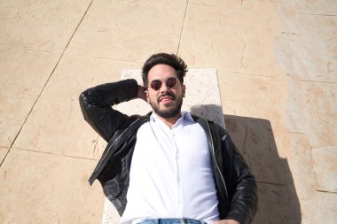 Sakallı, güneş gözlüklü, deri ceketli, beyaz gömlekli taş bir bankta yatan yakışıklı bir genç portresi. Güzelliği, modayı, eğilimi, huzuru, huzuru, huzuru kavra.