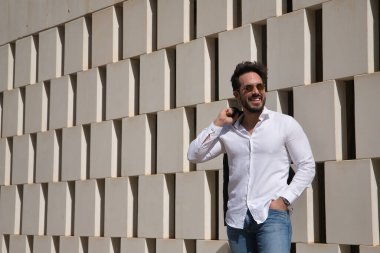 Sakallı, güneş gözlüklü, kılıçlı, deri ceketli, beyaz gömlekli ve kot pantolonlu, beyaz taş bloklardan oluşan duvarın yanında mutlu bir şekilde gülümseyen yakışıklı bir genç. Güzelliği, modayı, mutluluğu, kahkahayı kavra.