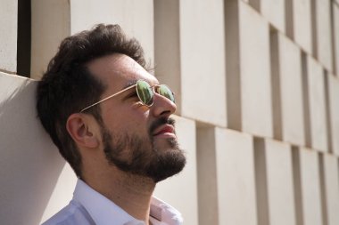 Sakallı, güneş gözlüklü ve beyaz gömlekli yakışıklı bir adamın portresi. Kafası beyaz taş bir duvara dayanıyor. Güzelliği, modayı, trendi, güneşi, yalnızlığı kavra.