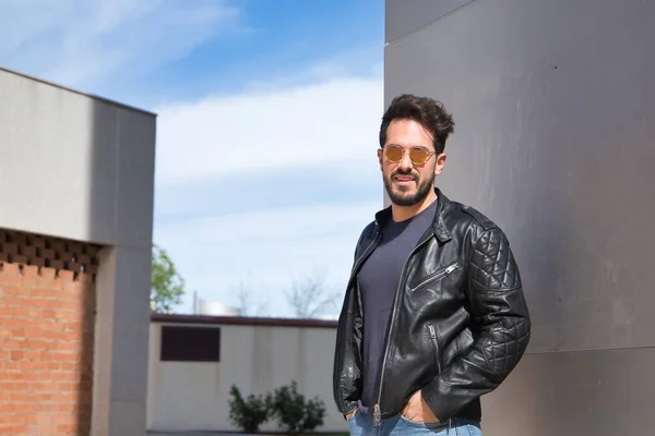 Handsome young man with beard, sunglasses, leather jacket and jeans, with his hands in his pockets and looking at the camera. Concept beauty, fashion, trend, modern.