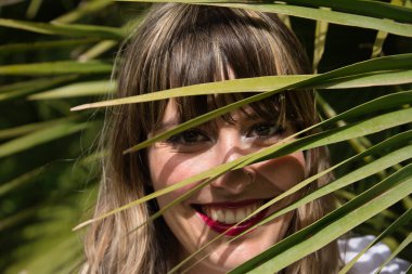 Yeşil bir palmiye ağacının yapraklarının arasından gülümseyen genç, güzel, sarışın bir kadının portresi. Güzellik kavramı, doğa, bahar, yaz, seyahat.