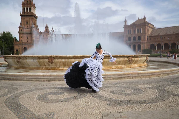 Young teenage woman in white dance suit with black polka dots and green carnations in her hair, dancing flamenco in front of a water fountain. Flamenco concept, dance, art, typical Spanish dance.