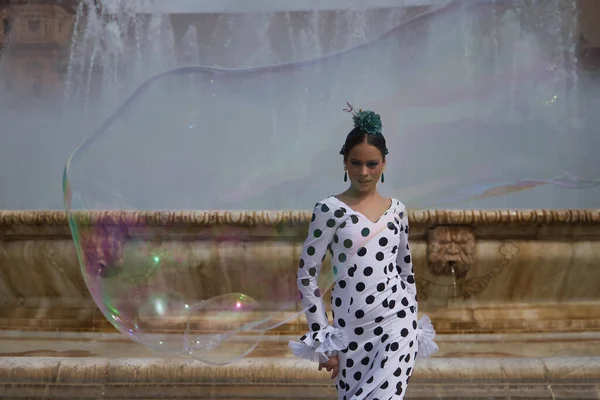 Young teenage woman in white suit with black polka dots, in front of a water fountain and seen through a large soap bubble. Flamenco concept, dance, art, typical Spanish dance, bubbles.