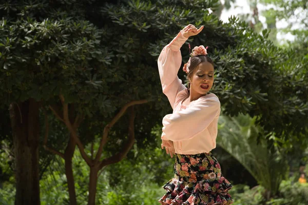 Young teenage woman in pink shirt, black skirt with flowers and pink carnations in her hair, dancing flamenco surrounded by greenery. Flamenco concept, dance, art, typical Spanish dance.
