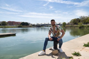 Genç ve yakışıklı bir adam, mavi gözlü, mükemmel gülüşlü, sakallı, şapkalı ve Hawaii gömlekli, nehir kıyısında bir iskelede oturuyor. Tatilleri, partileri, seyahatleri, seyahatleri.