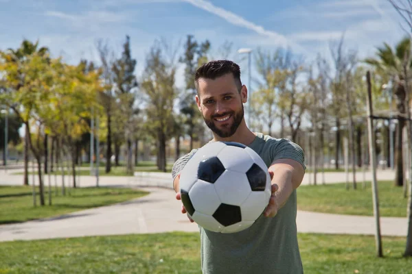 Young and handsome man, with a beard and green shirt, with blue eyes, perfect smile, offering a soccer ball, smiling. Concept sport, ball, football, world, competition.