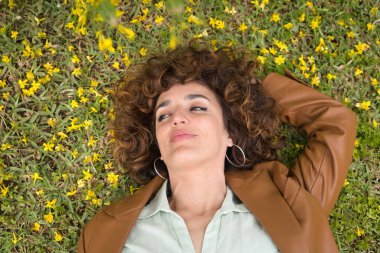 Sarı çiçeklerle çevrili çimlerin üzerinde kıvırcık kahverengi saçlı ve deri ceketli çekici olgun kadın portresi. Bahar konsepti, çiçekler, güzellik, saç stili, bukleler.