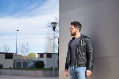Sakallı, güneş gözlüklü, deri ceketli ve kot pantolonlu, bir binanın duvarına yaslanmış, tasasız genç bir adam. Güzellik kavramı, moda, moda, modern.
