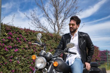 Sakallı, güneş gözlüklü, deri ceketli, beyaz gömlekli ve kot pantolonlu, büyük yer değiştiren motosikletli yakışıklı bir genç. Güzellik, moda, trend, motor, motosiklet, çıplak kavram.