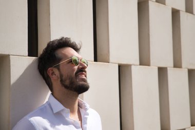 Sakallı, güneş gözlüklü, beyaz gömlekli beyaz taş bir duvara yaslanmış gülümseyen yakışıklı bir genç portresi. Güzelliği, modayı, trendi, güneşi, mutluluğu, barışı kavra.