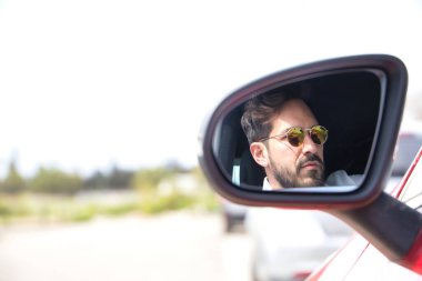 Kırmızı spor arabasının dikiz aynasında sakallı, güneş gözlüklü ve beyaz gömlekli yakışıklı genç adamın portresi yansıyor. Güzellik, moda, trend, lüks, motor, spor, kazanan, ayna.