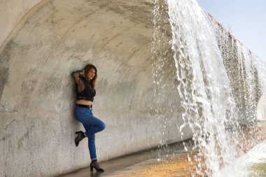 Çekici olgun bir kadın, şeffaf siyah gömlekli, kot pantolonlu ve topuklu, şehvetli ve kışkırtıcı bir tavırla şelalenin yanında poz veriyor. Olgunluk, güzellik, moda, baştan çıkarma, provokasyon kavramı.