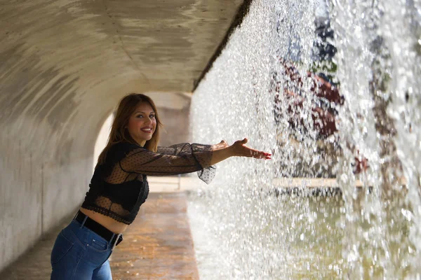 Attractive mature woman in transparent black shirt and jeans, touching the water falling from a waterfall. Concept maturity, beauty, fashion, water, happiness.