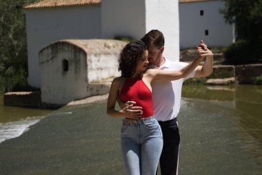 Taş bir zeminde, açık hava parkında, nehrin yanında dans eden çekici genç çift. Latin dansı konsepti, şehvetli, folklorik, kentsel.