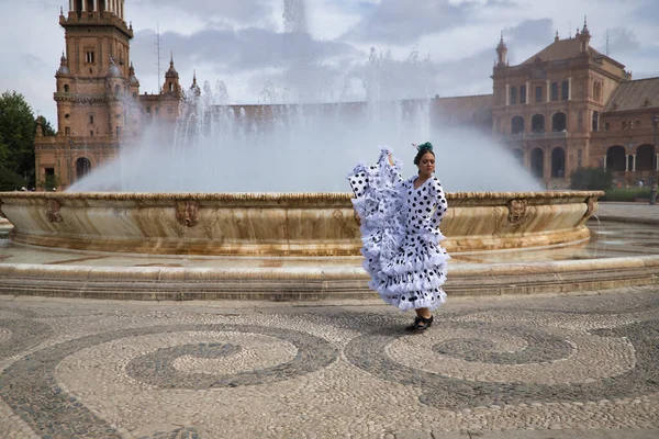 Young teenage woman in white dance suit with black polka dots and green carnations in her hair, dancing flamenco in front of a water fountain. Flamenco concept, dance, art, typical Spanish dance.