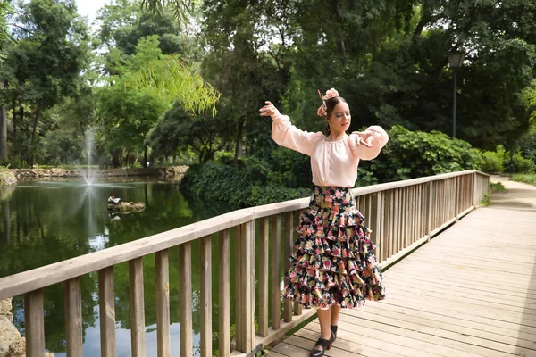 Young teenage woman in pink shirt, black skirt with flowers and pink carnations in her hair, dancing flamenco on wooden bridge by a lake. Flamenco concept, dance, art, typical Spanish dance.