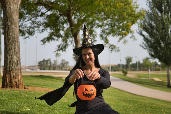 Woman in black witch suit and hat and showing a pumpkin she is holding in her hands, sitting on the grass in an outdoor park celebrating halloween. Autumn concept, trick or treat, party, pumpkin.