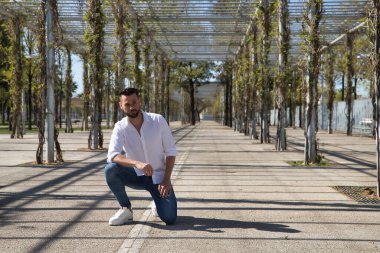 Sakallı, beyaz gömlekli, kot pantolonlu, dizlerinin üzerinde ve elleri bacaklarının üzerinde yakışıklı bir genç. Konsept moda, güzellik, modern moda, model.