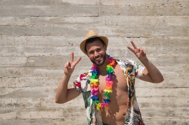 Young and handsome man, blue eyes, perfect smile, with beard, hat, open Hawaiian shirt and flower necklace and making a sign of victory on a gray background. Concept vacation, party, travel.
