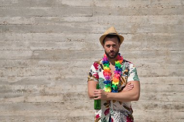 Young and handsome man, blue eyes, with beard, hat, Hawaiian shirt and flower necklace with arms crossed and a bottle of beer in his hand on a gray background. Concept holidays, party, trips, drink.