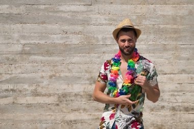 Young and handsome man, blue eyes, with beard, hat, Hawaiian shirt and flower necklace and holding a bottle of beer on a gray background. Concept vacation, party, travel, drink, copy space.
