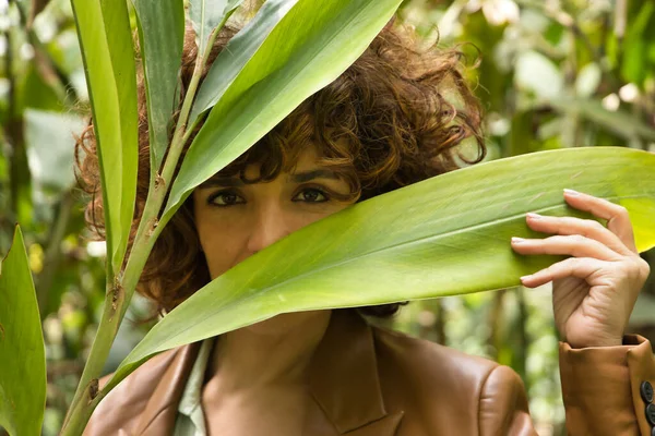 Attractive mature woman with curly brown hair, wearing brown leather jacket looking through large green leaves of a plant. Concept looks, happiness, hairstyle, plants, nature.